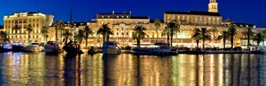 Een hotel aan het water met verschillende boten afgemeerd, een opvallende klokkentoren en palmbomen langs de promenade.
