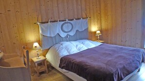 Um quarto acolhedor de madeira com uma cama, duas luminárias de cabeceira, uma poltrona e uma mesinha.