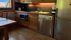 A compact kitchen with wooden cabinets, a stainless steel dishwasher, a microwave, and a refrigerator. - Comfortable apartment near center with balcony and parking (Samoëns)