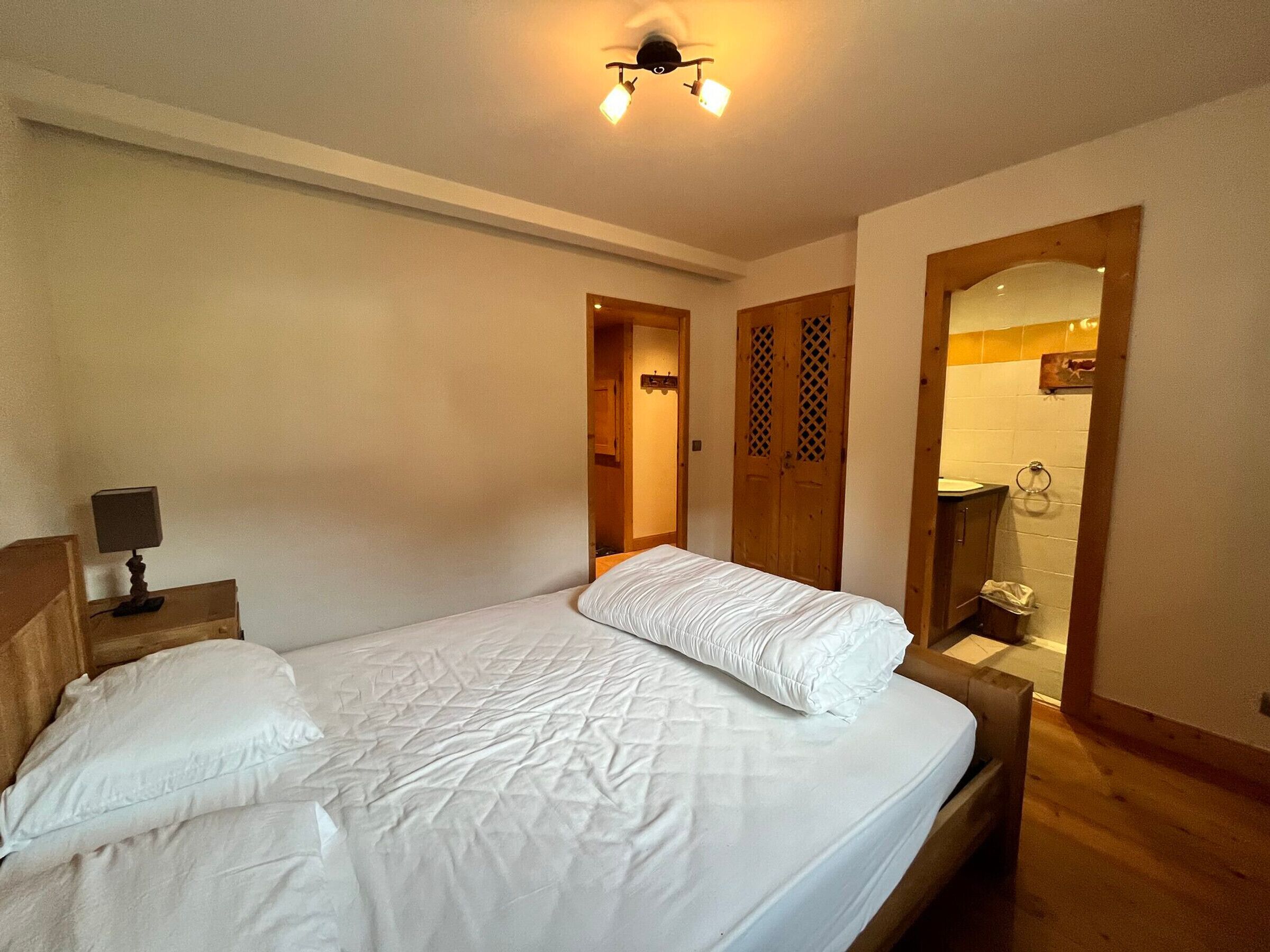 A bedroom with a bed, a bedside table with a lamp, a wooden wardrobe, and a bathroom visible through an open door.