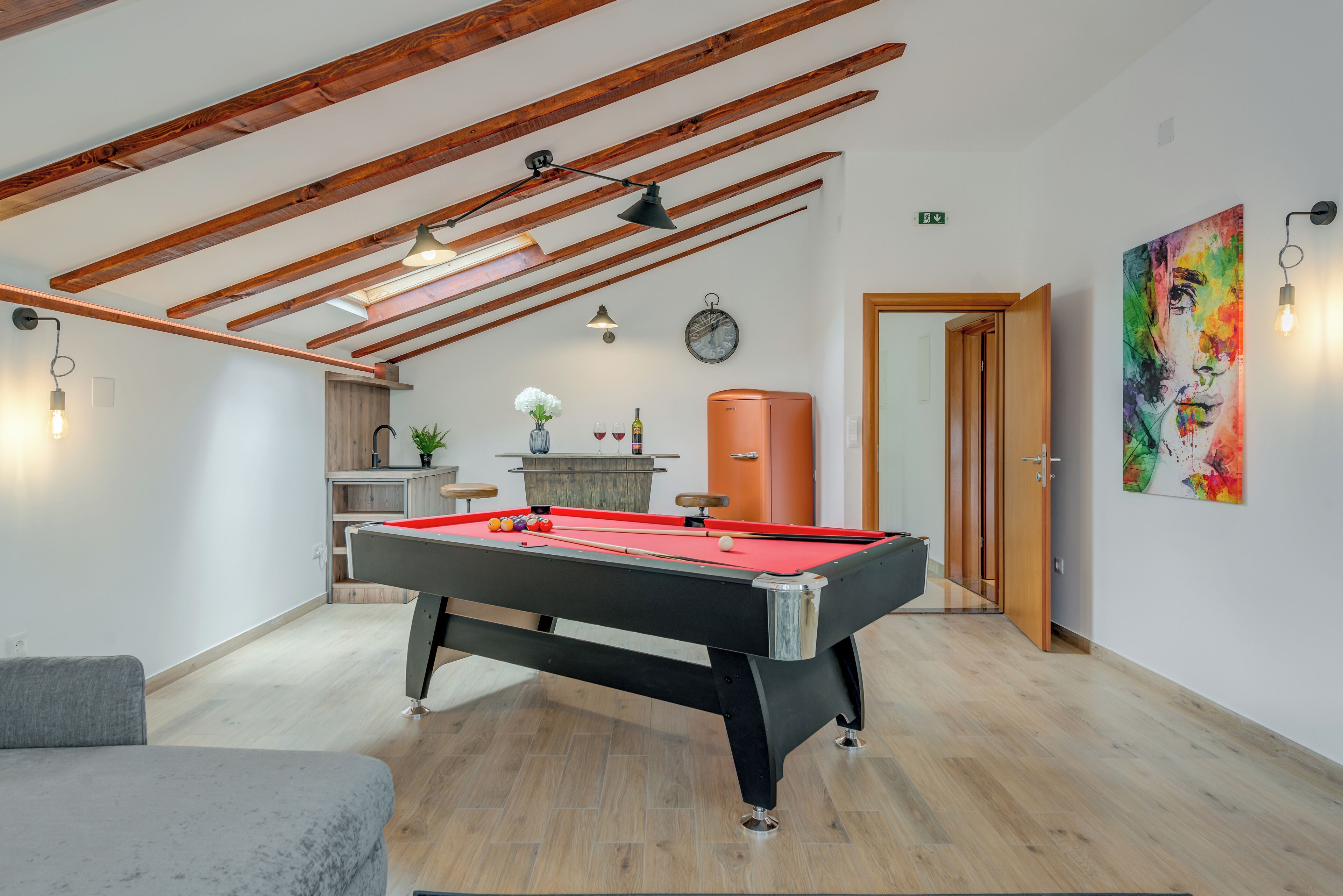 A pool table in a room with wooden beams on the ceiling, a clock on the wall, and a colorful painting.
