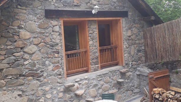 A stone cottage with a balcony, a small table with chairs, and a pile of firewood. - Chalet in a small village in the Hautes Pyrénées close to walks and hikes (Bareilles)