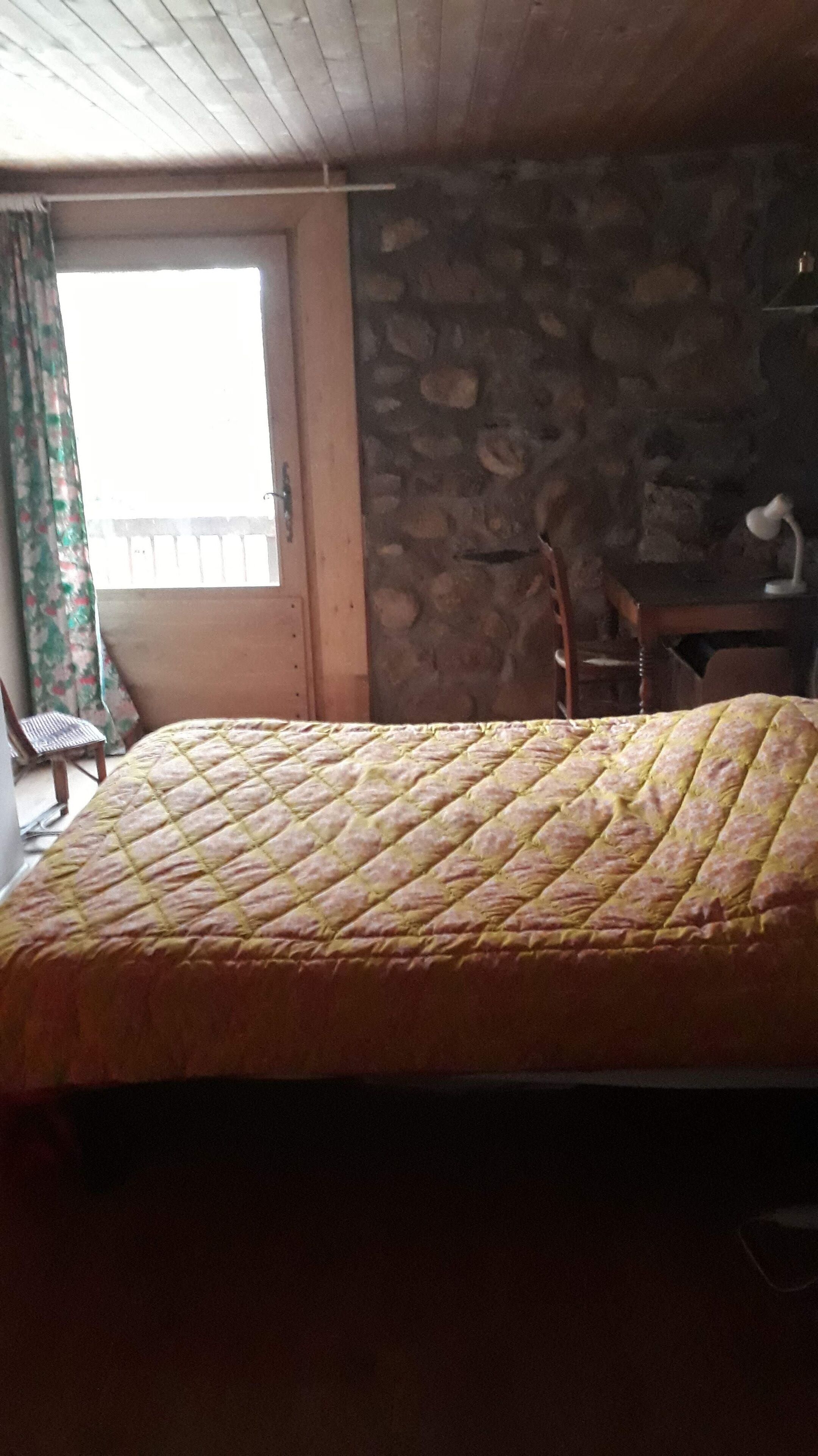 Ein Zimmer mit einem Bett, einem Sessel, einem Schreibtisch, einem Fenster und einer Steinwand.
