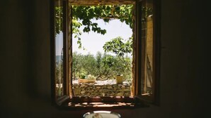 Um quarto rústico com parede de pedra, uma mesa pequena com um prato e uma janela com vista para um jardim com árvores e um banco.