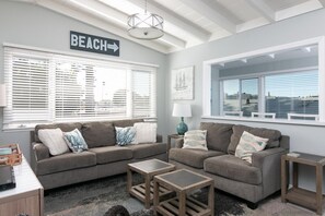 A living room with a gray sofa set, two side tables, a lamp, and a view of the beach through large windows. - Beach-block Charming Upstairs Duplex (Ventura)