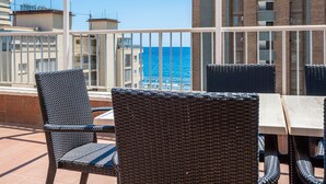 Ein Balkon mit Korbstühlen und einem Tisch, mit Blick auf ein Gebäude und das Meer.