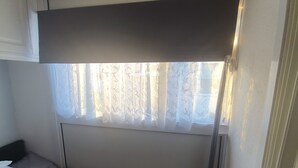 A small, compact bedroom with a single bed, a window with sheer curtains, and a door to the right.