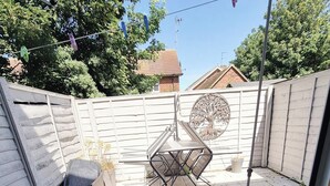 Une cour arrière clôturée avec une table et des chaises en métal, une découpe décorative en forme d’arbre sur la clôture et une boîte de rangement noire.
