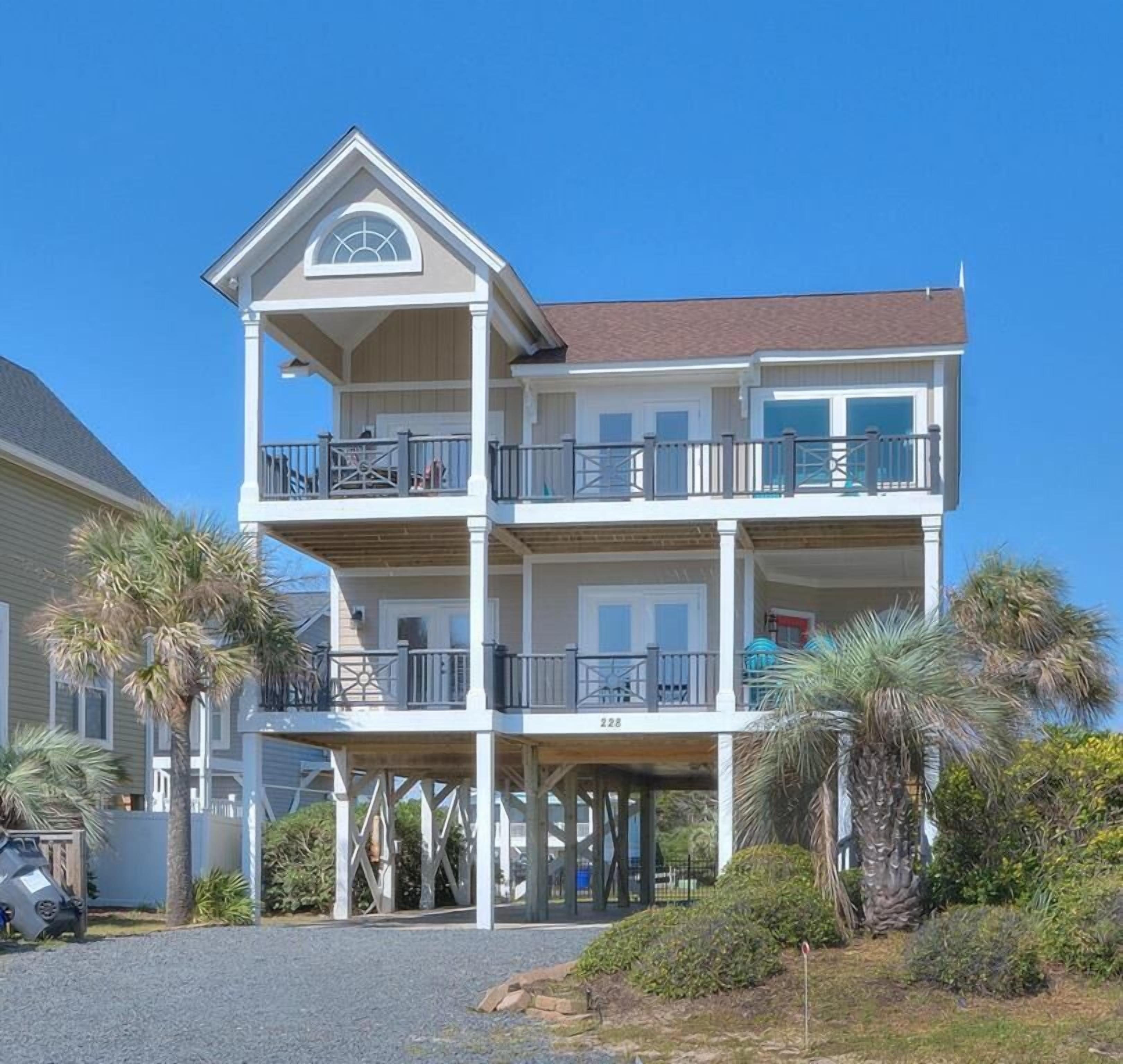 A multi-story beachfront property with a balcony on each floor, a covered front porch, and a clear view of the ocean.