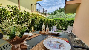 Un área de comedor al aire libre con una mesa dispuesta para dos personas, rodeada de follaje y con vista a un edificio residencial.
