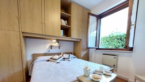 Un dormitorio con cama, armario de madera, mesita de noche con lámpara y ventana con vista a follaje.