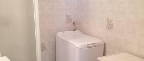 A bathroom with a white washing machine, a toilet, and a small shelf unit.
