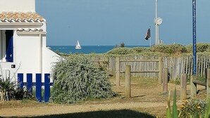 Edificio costero con fachada blanca y detalles azules, valla de madera y vista despejada al mar con un velero.