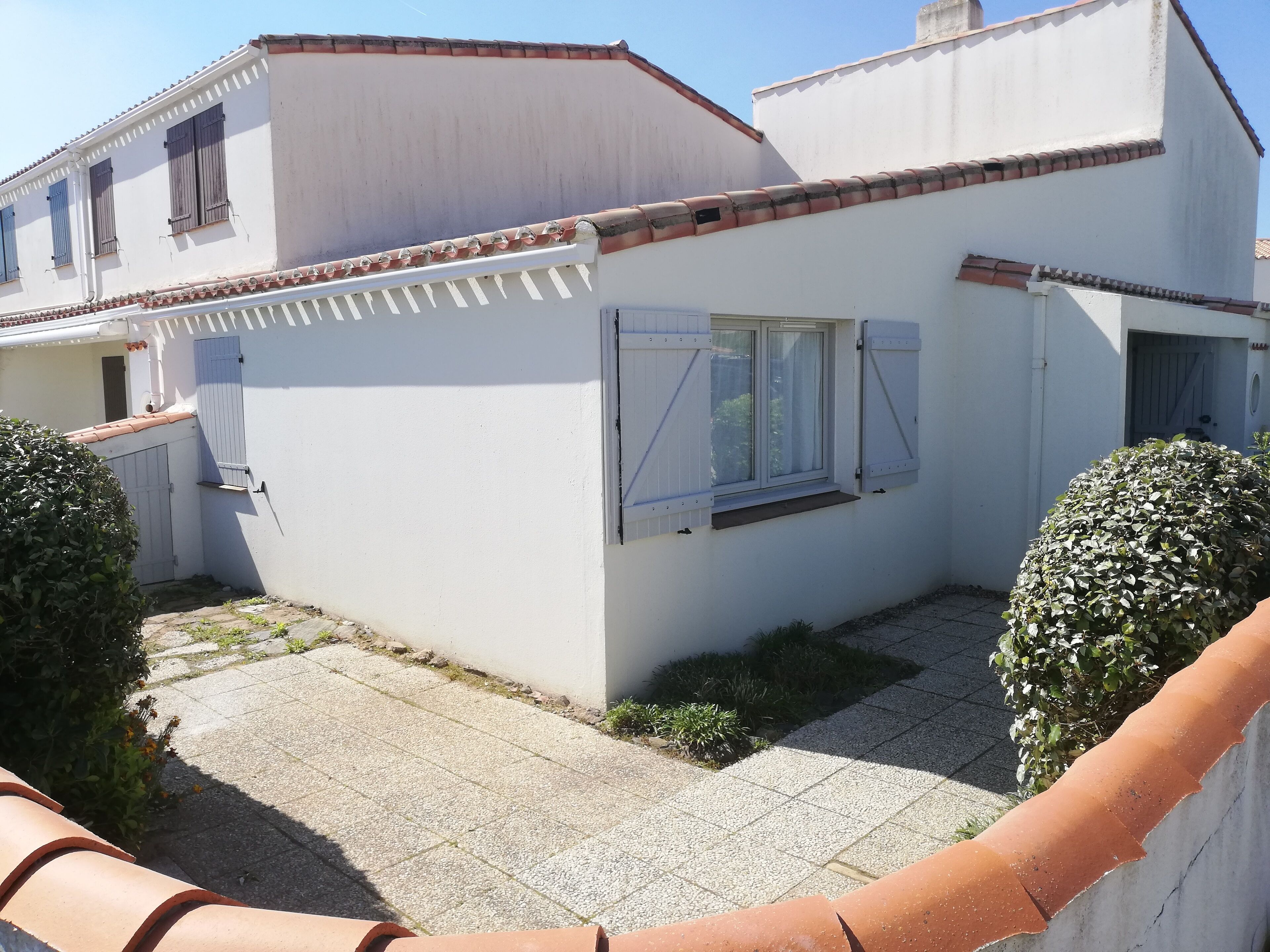 A white building with a tiled roof, a window with shutters, and a small garden area with a hedge.