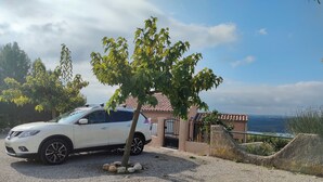 Un VUS blanc est garé sur une allée de gravier, à proximité d’un arbre, d’une maison avec un toit en tuiles, et offre une vue sur la mer.