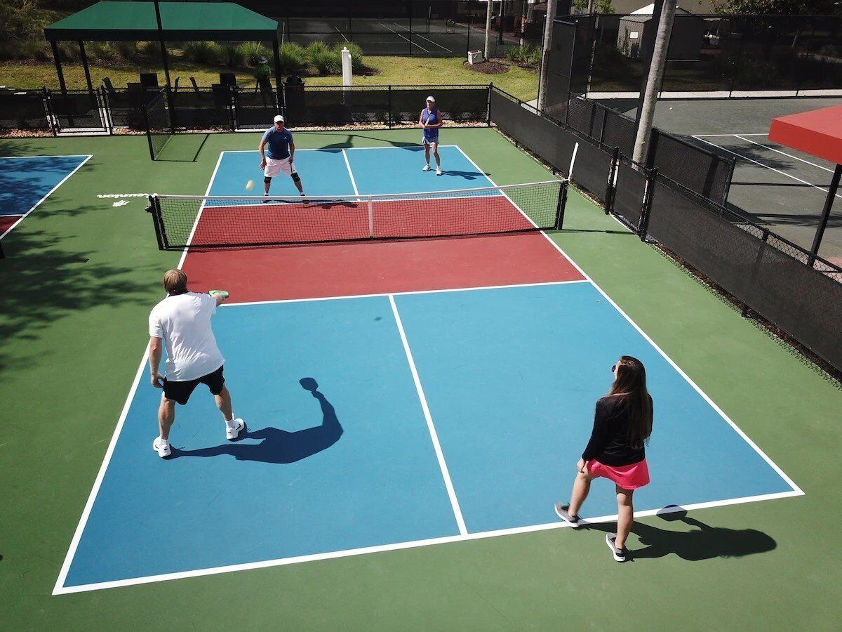 Camp de tennis amb quatre jugadors, una xarxa i una tanca verda al voltant del camp.