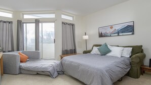 A bedroom with a bed, a sofa, a nightstand, a lamp, and a painting on the wall. - 208 Village Gate Studio Bedroom Condo by RedAwning (Warren)