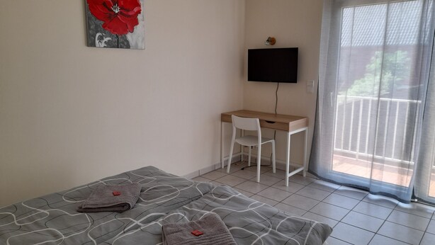 Quarto com cama, escrivaninha, cadeira, televisão e janela com cortinas.