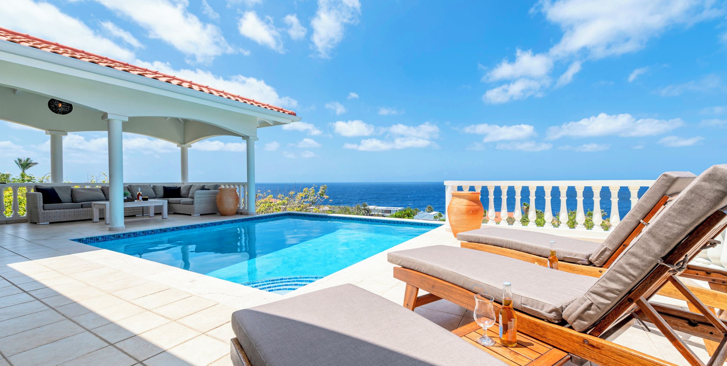 A pool area with lounge chairs, a covered seating area, and a view of the ocean.