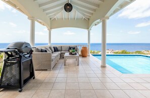 A covered outdoor seating area with a grill, a sofa, a coffee table, and a view of the ocean.