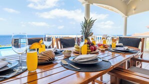 A dining table set for two with a view of the ocean, a pineapple, and various fruits and beverages.