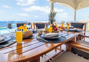 Ein Esstisch für zwei Personen mit Blick auf das Meer, eine Ananas und verschiedene Früchte sowie Getränke.