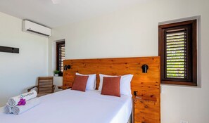 A hotel room with a wooden headboard, a bed with white bedding, two brown pillows, a rolled towel, a wooden chair, and a small table with a plant.