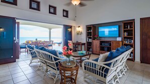 A spacious living room with a large TV, wicker furniture, and a view of the sea. - Villa Azure Rose - Coral Estate (Curaçao)