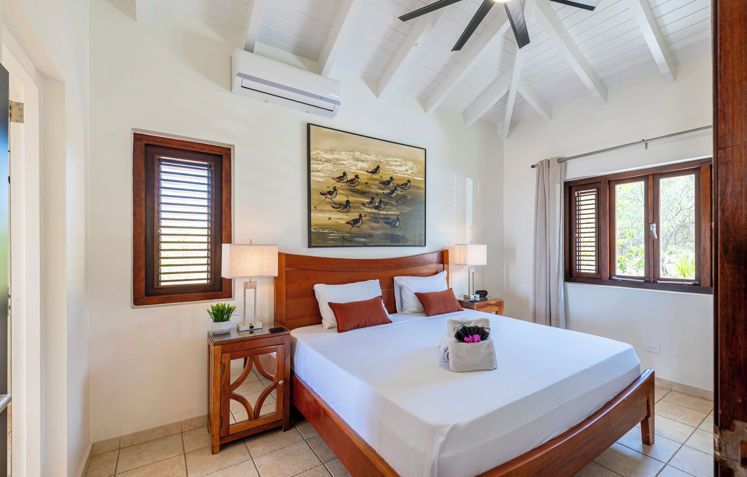 A hotel room with a wooden bed, a painting on the wall, a window with blinds, and a ceiling fan.