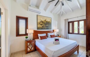 A hotel room with a wooden bed, a painting on the wall, a window with blinds, and a ceiling fan. - Villa Sundara - Coral Estate (Curaçao)