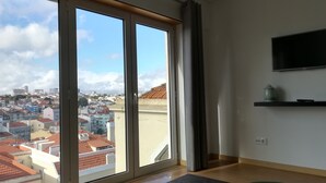 A spacious room with large windows offering a city view, a flat-screen TV mounted on the wall, and a small shelf with decorative items. - Lisbon Grand View (Lisboa)