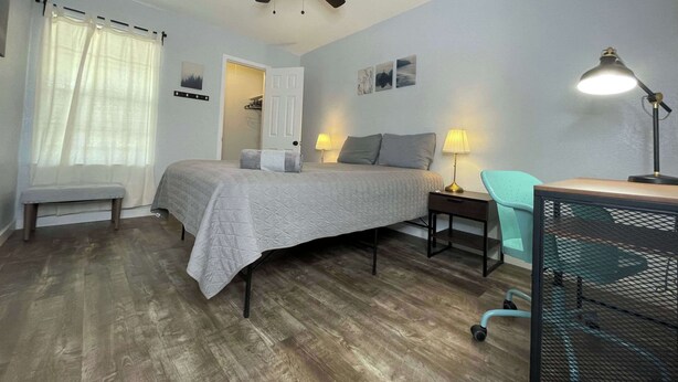 Quarto com cama, mesas de cabeceira, escrivaninha, cadeira, ventilador de teto e janela com cortinas.