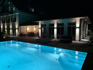 Ein Schwimmbecken mit einer eingelassenen Sitzgruppe, eine überdachte Terrasse mit Tischen und Stühlen sowie ein großes Haus mit zahlreichen Fenstern und Türen.