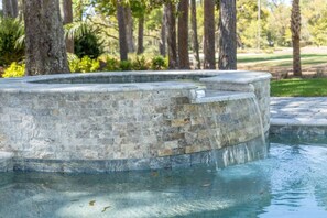 A stone fountain with water cascading down its sides, surrounded by trees and a grassy area.