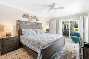 A bedroom with a bed, a nightstand, a lamp, a ceiling fan, and a view of the outdoors.