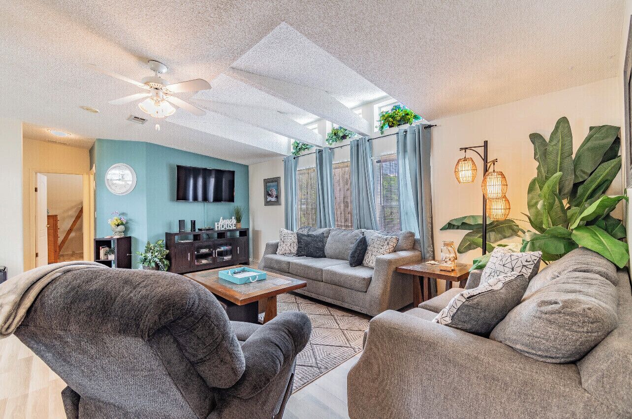 Ein gemütliches Wohnzimmer mit einer Couch, Sesseln, einem Couchtisch und einem Fernseher. Es gibt einen Deckenventilator, ein Fenster mit Vorhängen und eine Pflanze.
