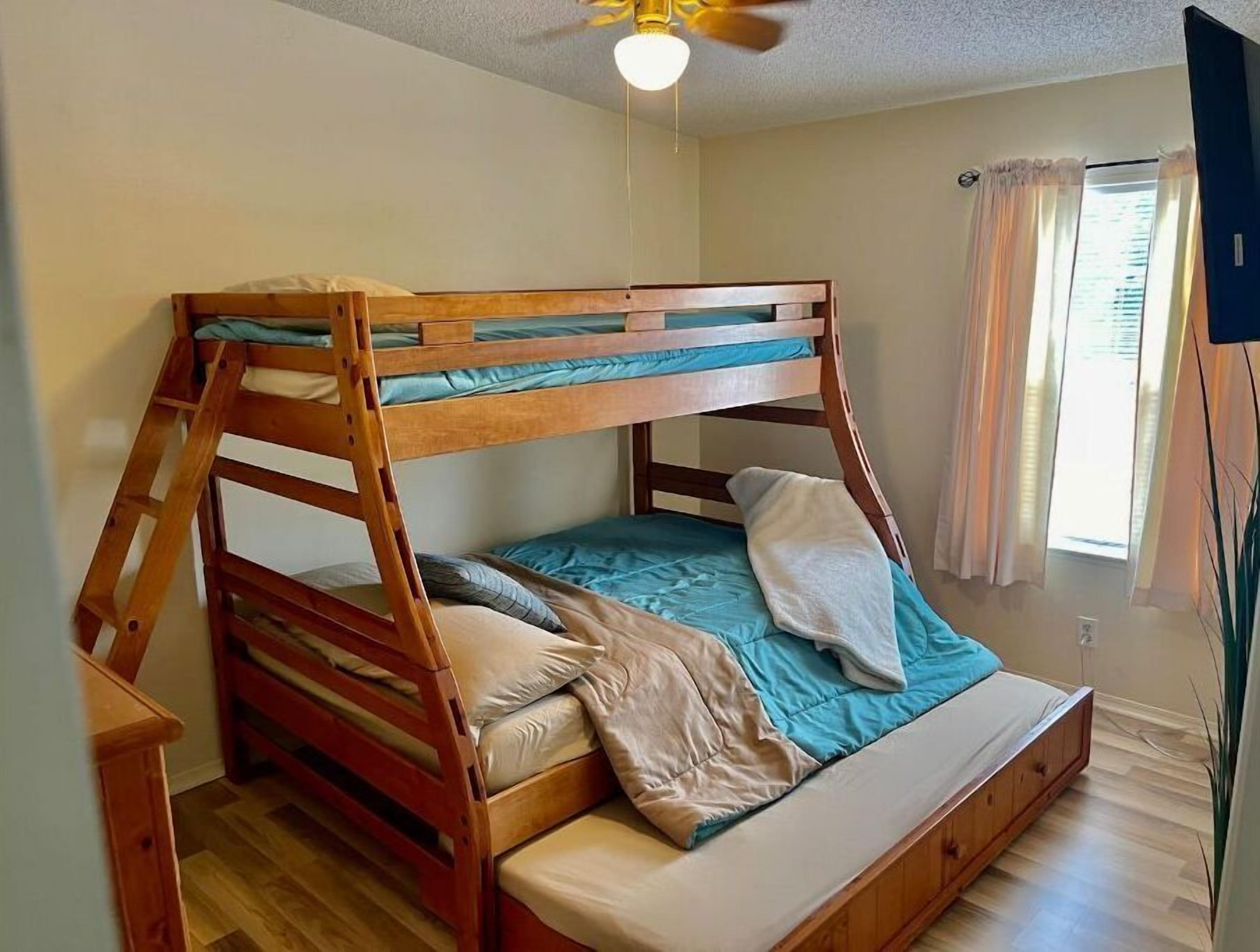 Una cama litera con estructura de madera, un ventilador de techo y una ventana con cortinas.