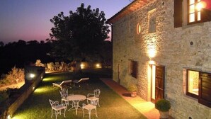 A stone building with a well-lit outdoor seating area, including a table and chairs, and a small garden. - Podere Lucignano Secondo - La Mangiatoia (Gaiole in Chianti)