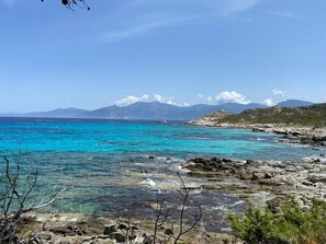 Una costa rocosa con agua turquesa cristalina y montañas distantes bajo un cielo despejado.