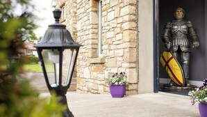 L’entrée d’un bâtiment en pierre est surmontée d’une statue d’un chevalier tenant un bouclier. Deux pots de fleurs violettes sont posés sur le sol.