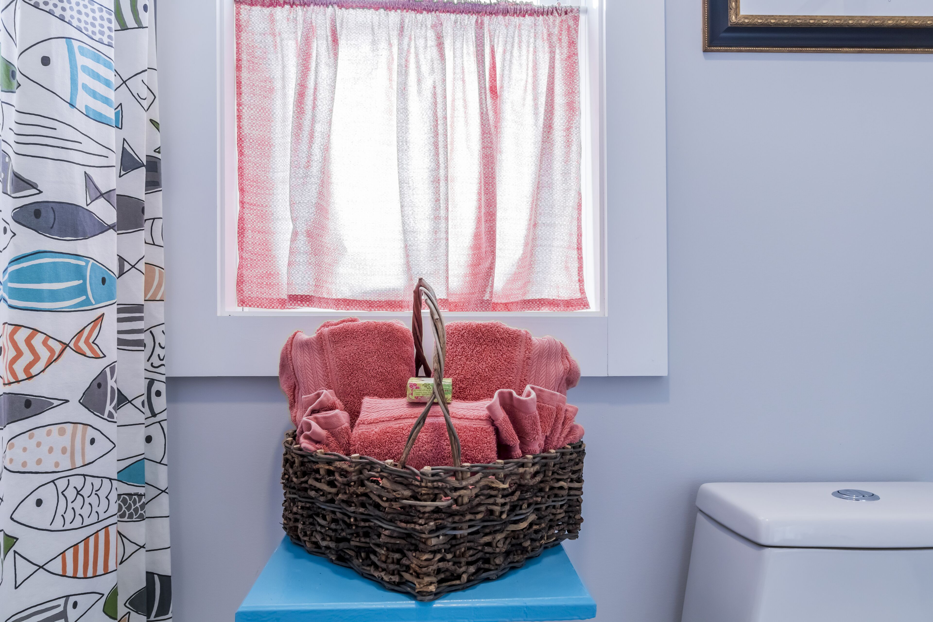A bathroom with a blue sink, a white toilet, a wicker basket with pink towels, and a window with pink curtains.