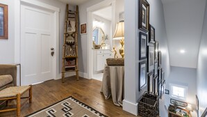 A hallway with a wooden floor, a chair, a ladder, and a table with a lamp.