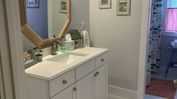 Une salle de bain avec un meuble-lavabo blanc, un grand miroir et un évier. On y trouve des tableaux encadrés accrochés au mur, un porte-serviettes et un rideau de douche orné d’un motif de poissons.