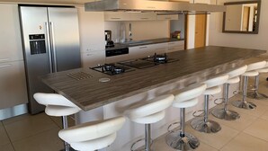 A modern kitchen with a central island, bar stools, stainless steel appliances, and a large window. - Villa Contemporaine D'exception à 5 min de Propriano (viggianello)