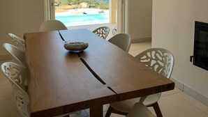 A dining area with a wooden table and white chairs, a bowl of fruit, and a view of the ocean and buildings. - Villa Contemporaine D'exception à 5 min de Propriano (viggianello)