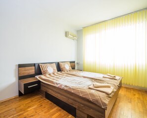 A bedroom with a wooden bed, a nightstand, a built-in drawer, a wall-mounted air conditioner, and a window with vertical blinds. - Aleks Classic (Burgas)