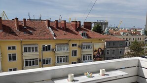 Un balcón con una silla blanca, una mesita con una taza y vistas a un paisaje urbano con edificios de tejas rojas.