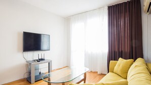 Una sala de estar moderna con una mesa de centro de cristal, un sofá amarillo, un televisor de pantalla plana sobre un mueble bajo y una ventana con cortinas.