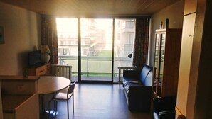A compact living space with a dining table, a sofa, and a kitchenette. A large sliding glass door leads to an outdoor area with a view of a building and a green space. - Studio vue sur mer Avec Piscine Interieure (La Panne)