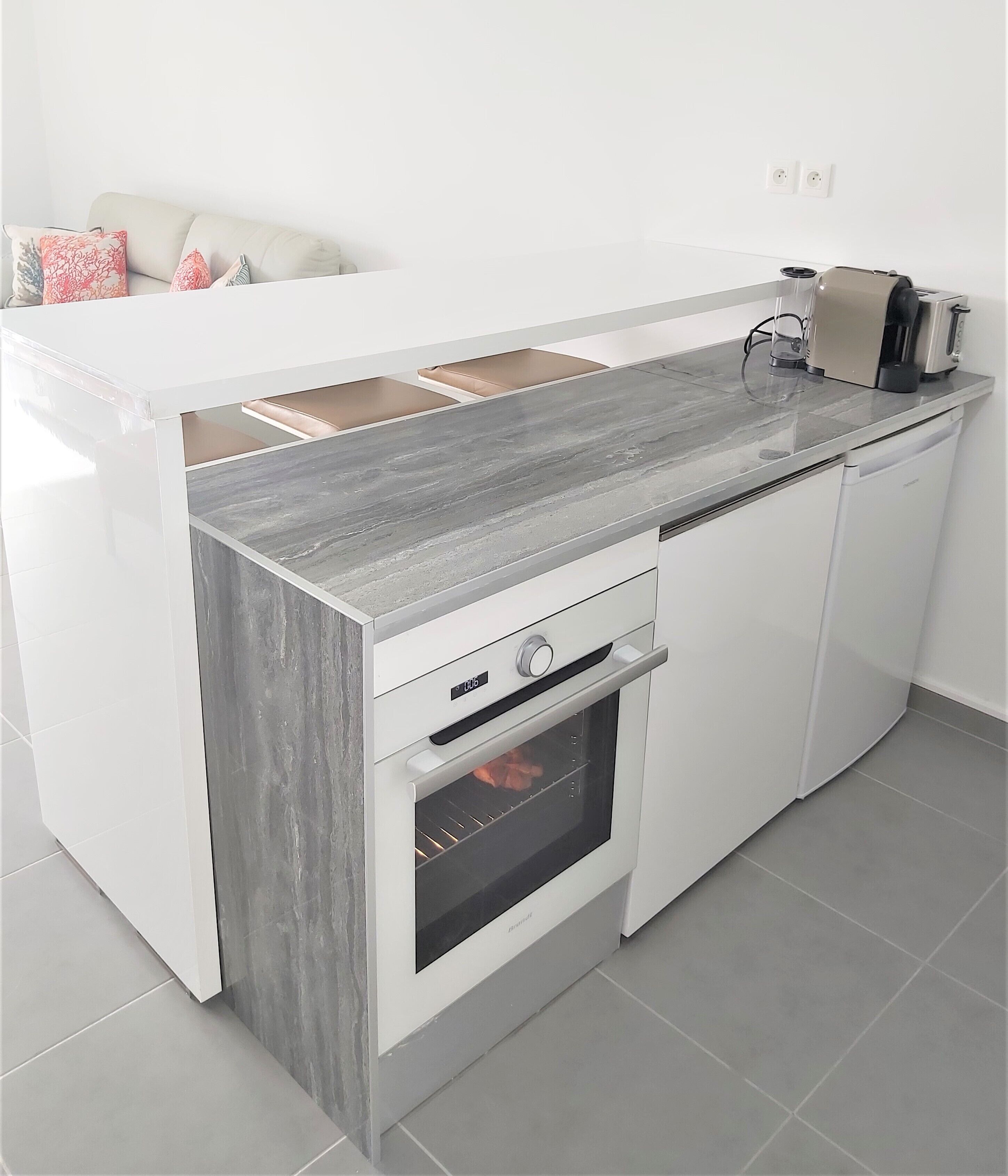 Une cuisine compacte dotée d’un four encastré, d’un plan de travail à la finition imitant le marbre gris et de meubles blancs.
